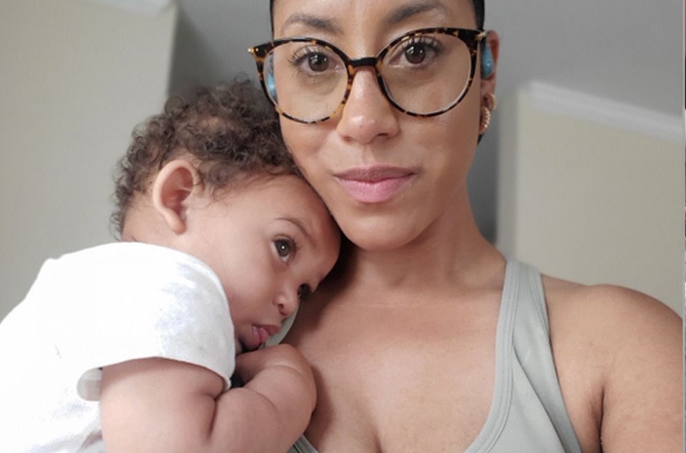 Patience in gray tank top and glasses, holding infant on shoulder, both looking away.