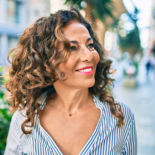 Una mujer sonriente con cabello rizado está parada afuera mirando hacia un lado con una expresión feliz.