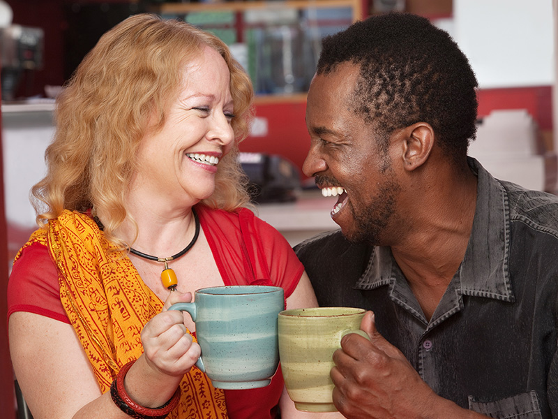 A smiling couple is sitting together, laughing and holding mugs. 