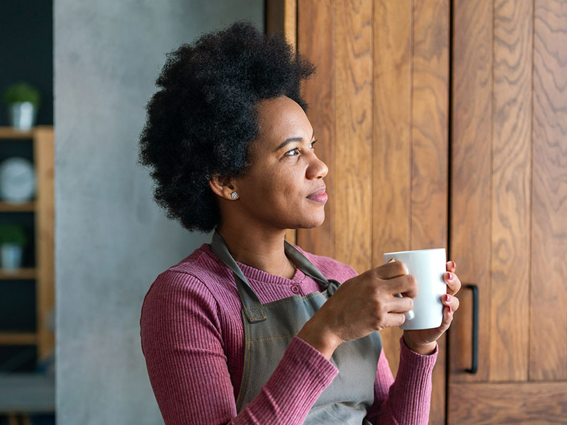 A woman wearing an apron sitting in a chair, holding a mug and looking out the window. 