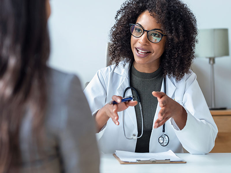 A doctor with curly hair and glasses, with a stethoscope around her neck, is sitting at a desk, engaging in a conversation with a patient while holding a pen and gesturing with her hands.