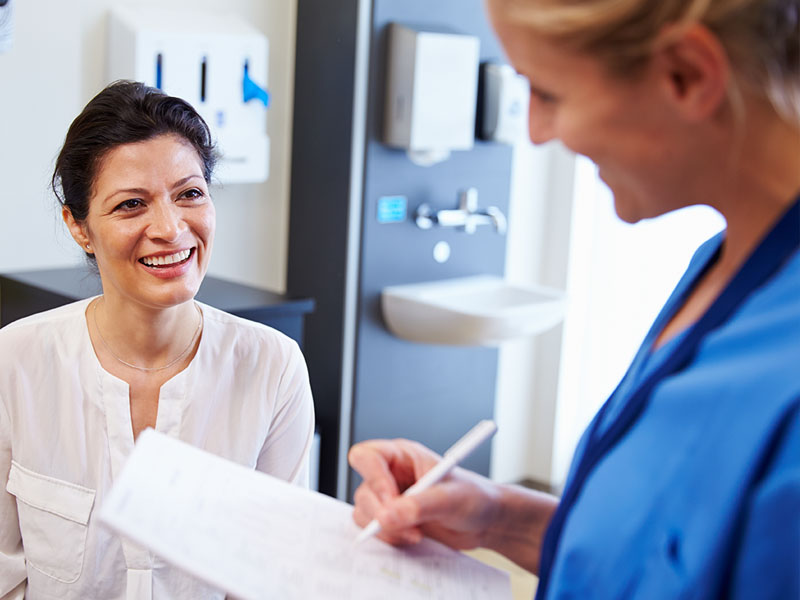 A woman is smiling while sitting in a medical office, as a healthcare professional in blue scrubs is writing on a piece of paper. 